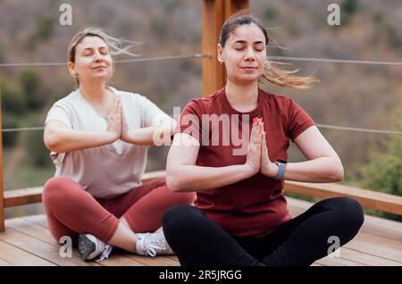 Zwei junge Frauen trainieren Asana-Yoga im Park. Lächelnde Mädchen sitzen in Lotusposition. Weibliche Freunde meditieren auf einer offenen Holzplattform. Gesund Stockfoto