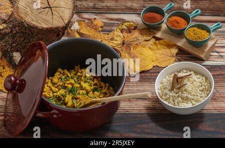 Ein Tontopf mit Hühnchen-Curry drinnen, auf der rechten Seite eine Tasse weißen Reis mit Zimt und im Hintergrund ein Baumstamm, trockene Blätter und drei Schüsseln wi Stockfoto