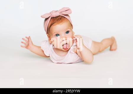 Porträt eines niedlichen, rothaarigen Babys auf dem Bauch, weißer Hintergrund, mit pinkfarbenem Körper und Stirnband Stockfoto