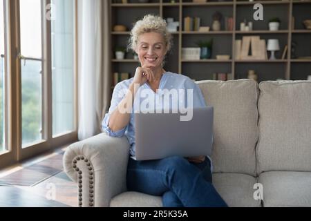 Glückliche, attraktive, reife Dame, die den Laptop zu Hause hält Stockfoto