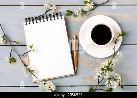 Künstlerische Komposition mit geöffnetem Notizblock und Stift, Tasse schwarzen Kaffee, Zweig weißer Kirschblüten auf blauem Holztisch. Festlicher Büroschreibtisch Stockfoto