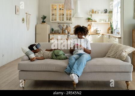 Die vielbeschäftigte afroamerikanische Mutter benutzt ihr Smartphone, während ihr kleiner Sohn zu Hause mit einer VR-Brille einen Film anschaut Stockfoto