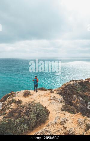 Ein Abenteurer mit einem Rucksack steht am Rand einer Klippe und beobachtet die wunderschöne Gruppe von gelb-goldenen Felsen von Punta de la Piedad in Lagos, im Stockfoto