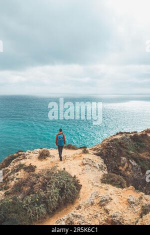 Ein Abenteurer mit einem Rucksack steht am Rand einer Klippe und beobachtet die wunderschöne Gruppe von gelb-goldenen Felsen von Punta de la Piedad in Lagos, im Stockfoto