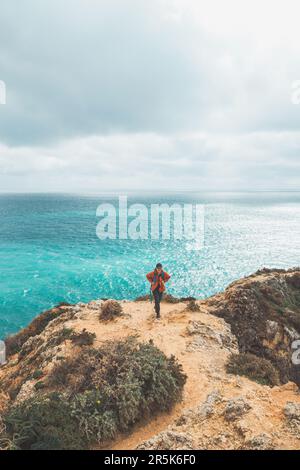 Ein Abenteurer mit einem Rucksack steht am Rand einer Klippe und beobachtet die wunderschöne Gruppe von gelb-goldenen Felsen von Punta de la Piedad in Lagos, im Stockfoto