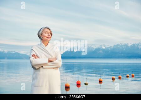 Außenporträt einer wunderschönen Frau mittleren Alters, die sich am Wintersee posiert und einen weißen Mantel trägt Stockfoto