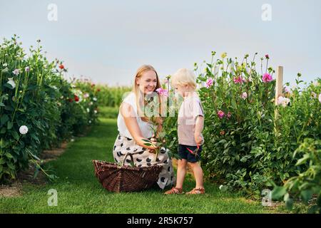 Glückliche junge Mutter und kleiner Junge, die in einem wunderschönen Blumengarten arbeiten, Blumen pflücken, Familienleben Stockfoto