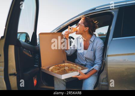 Eine Frau mittleren Alters isst gerade italienische Pizza, sitzt auf dem Fahrersitz während der Essenspause und genießt den Sonnenuntergang. Autofahrt, Fast Food je Stockfoto