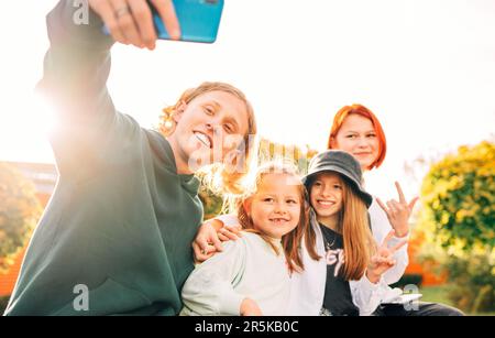 Porträts von drei lächelnden Schwestern und einem Bruder des Teenagers, der mit einer modernen Smartphone-Kamera Selfie-Portraits aufnahm. Unvorsichtiger junger Teenholz und kindheitszeitalter tim Stockfoto