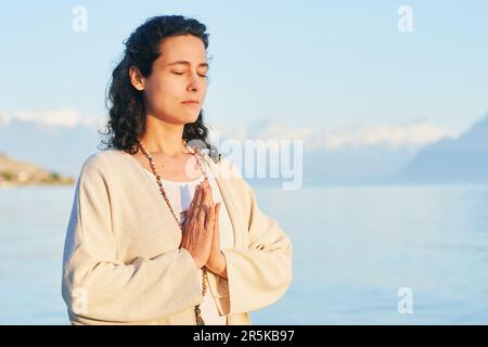 Wunderschöne spirituelle Frau, die am See meditiert, beige Kleidung trägt Stockfoto