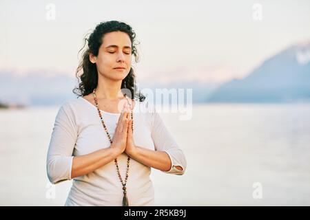 Wunderschöne spirituelle Frau, die am See meditiert, weiße Kleidung trägt Stockfoto