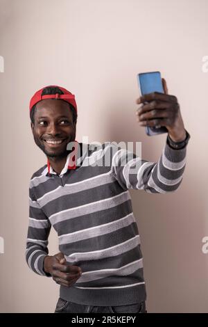 Junger Mann mit trendy Outfit und Mütze, der Selfies mit dem Smartphone in einer sauberen Studioumgebung macht. Perfekt für moderne Lifestyle- und Technikthemen.“ Stockfoto