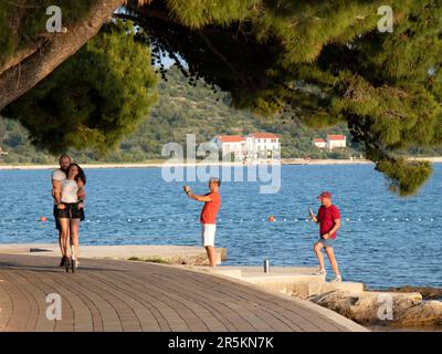 Vodice, Kroatien - 30. Mai 2023: Promenade mit Kiefern am Meer und Menschen, die auf einem elektrischen Roller unterwegs sind, bei Sonnenuntergang Stockfoto