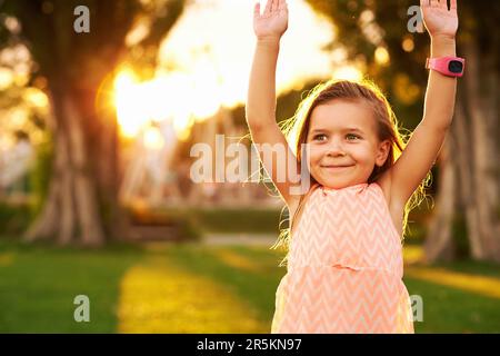 Außenporträt eines bezaubernden 3- oder 4-jährigen Mädchens, das im Sommerpark spielt, erhobene Arme, eine Smartwatch trägt, wunderschönes Sonnenlicht auf einem hellgrünen Rasen Stockfoto