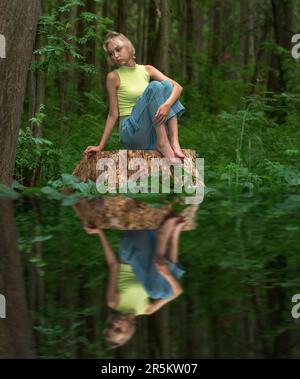 Teenager sitzt auf einem Baumstumpf in einem Naturwaldpark mit einem traurigen und durchdachten Look, der sich im Wasser widerspiegelt Stockfoto