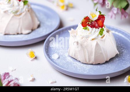 Zwei kleine pavlova-Minikuchen aus Baiser, dekoriert mit frischen Erdbeeren auf einem violetten Pastellteller. Helle Szene mit Blumen als Dekoration. Stockfoto