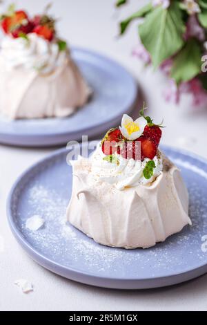 Zwei kleine pavlova-Minikuchen aus Baiser, dekoriert mit frischen Erdbeeren auf einem violetten Pastellteller. Helle Szene mit Blumen als Dekoration. Stockfoto