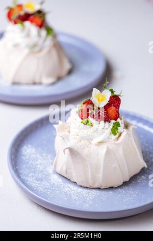 Zwei kleine pavlova-Minikuchen aus Baiser, dekoriert mit frischen Erdbeeren auf einem violetten Pastellteller. Helle Szene mit Blumen als Dekoration. Stockfoto