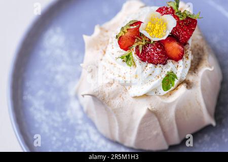 Zwei kleine pavlova-Minikuchen aus Baiser, dekoriert mit frischen Erdbeeren auf einem violetten Pastellteller. Helle Szene mit Blumen als Dekoration. Stockfoto