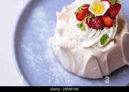 Zwei kleine pavlova-Minikuchen aus Baiser, dekoriert mit frischen Erdbeeren auf einem violetten Pastellteller. Helle Szene mit Blumen als Dekoration. Stockfoto