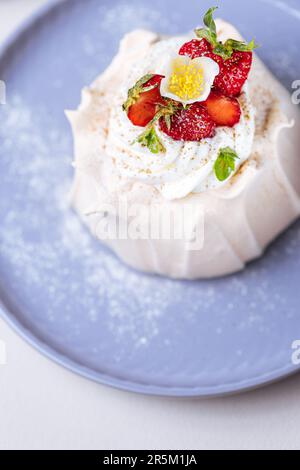 Zwei kleine pavlova-Minikuchen aus Baiser, dekoriert mit frischen Erdbeeren auf einem violetten Pastellteller. Helle Szene mit Blumen als Dekoration. Stockfoto