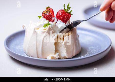 Zwei kleine pavlova-Minikuchen aus Baiser, dekoriert mit frischen Erdbeeren auf einem violetten Pastellteller. Helle Szene mit Blumen als Dekoration. Stockfoto