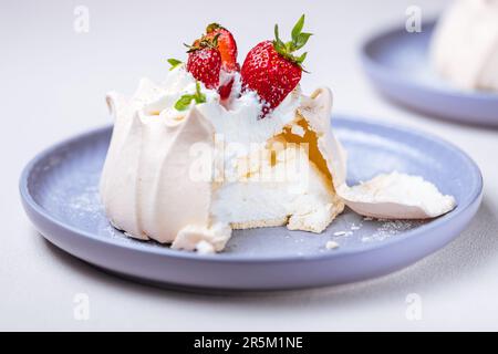 Zwei kleine pavlova-Minikuchen aus Baiser, dekoriert mit frischen Erdbeeren auf einem violetten Pastellteller. Helle Szene mit Blumen als Dekoration. Stockfoto