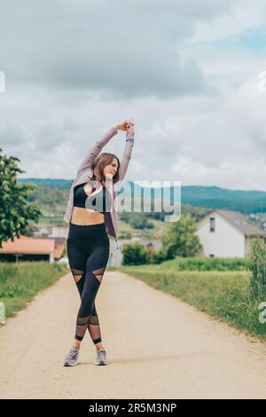 Außenporträt einer jungen, wunderschön sitzenden Frau in schwarzer Aktivbekleidung, Stretching von Sportlermodellen, Sportmode Stockfoto