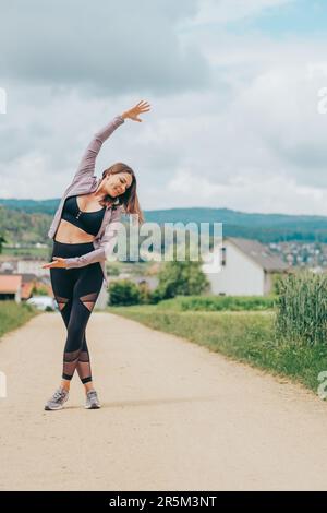 Außenporträt einer jungen, wunderschön sitzenden Frau in schwarzer Aktivbekleidung, Stretching von Sportlermodellen, Sportmode Stockfoto