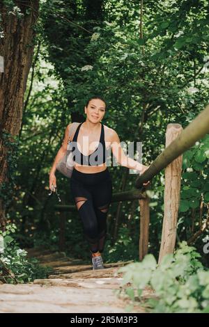 Fit für eine junge Frau, die im Wald wandert, die Treppe hochklettert, schwarze Aktivkleidung und einen Rucksack trägt Stockfoto