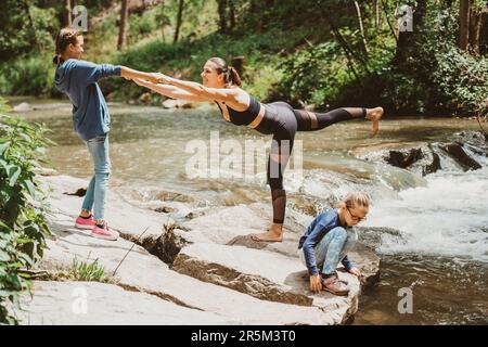 Junge, fitte Mutter, die Yoga in der Natur mit ihren Töchtern, ihrer sportlichen Familie und ihrem Sportflugzeug praktiziert Stockfoto