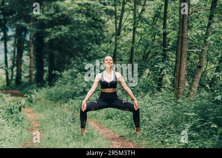 Junge Frau, die Yoga im Sommerwald praktiziert, trägt schwarze Leggings und ein Oberteil Stockfoto