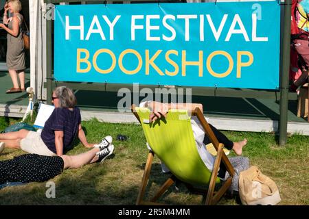 Die Leute entspannen sich in Liegestühlen und sitzen draußen auf dem Gras am Hay Festival Bookshop Schild am Hay on Wye in Sunshine 2023 Wales UK KATHY DEWITT Stockfoto