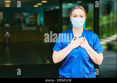 Junge weibliche Peeling-Krankenschwester trägt blaue Uniform und Gesichtsmaske, steht im Krankenhauseingang Stockfoto