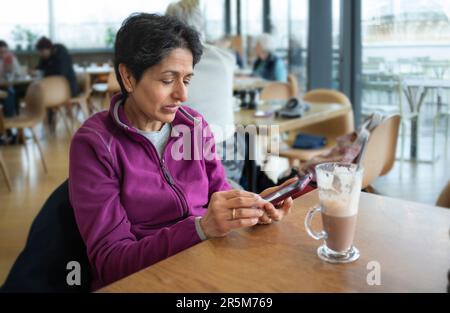 Eine indische Asiatin mittleren Alters, die allein in einem Café sitzt und ein Handy benutzt, Oxford, Großbritannien Stockfoto
