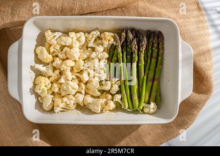 Frische rohe Blumenkohlblüten aus biologischem Anbau und grüner Spargel in einer Backform, Nahaufnahme auf dem Küchentisch, fertig zum Backen, Blick von oben Stockfoto