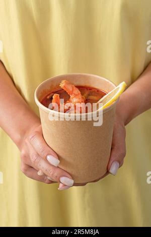 Traditionelle asiatische Suppe Tom Yum Kung in Einwegbecher Kraftpapier. Suppe zum Mitnehmen, Essenslieferung. Essen auf gelbem Hintergrund mitnehmen. Stockfoto