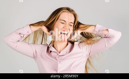 Porträt eines lustigen blonden Mädchens, das Zunge zeigt. Glückliche Frau mit langen Haaren und geschlossenen Augen. Lächelnde Frau mit unordentlichen Haaren in den Händen. Weibliches Modell mit Stockfoto