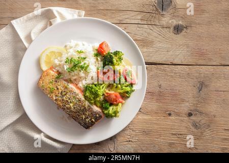 Knusprig gebratenes Lachsfilet mit Gemüse aus Brokkoli und Tomaten, Reis, Petersilie und Zitronenschale auf einem rustikalen Holztisch, Kopierbereich, Blick von oben Stockfoto