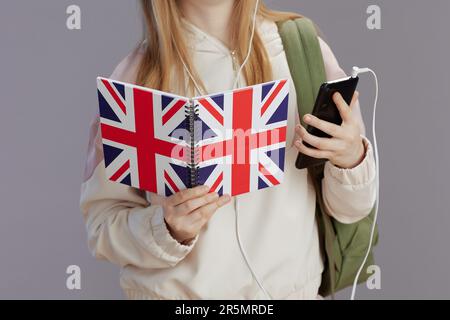 Nahaufnahme eines Mädchens mit Rucksack, arbeitsbuch mit englischer Flagge, Kopfhörer und Smartphone gegen Grau. Stockfoto