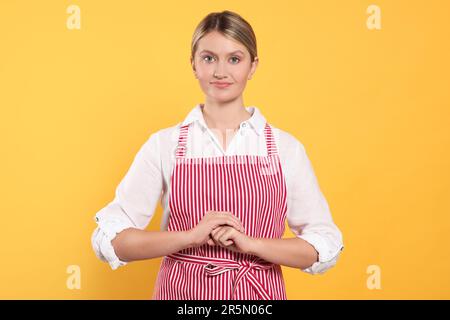 Wunderschöne junge Frau in einer sauberen gestreiften Schürze auf orangefarbenem Hintergrund Stockfoto