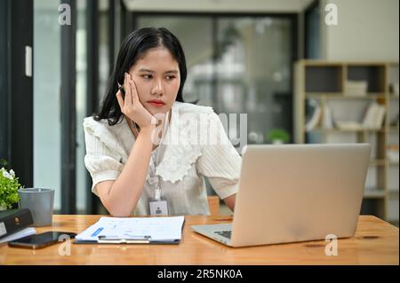 Müde und gelangweilte junge asiatische Büroangestellte arbeiten an ihrem Projekt an ihrem Schreibtisch im Büro. Stockfoto
