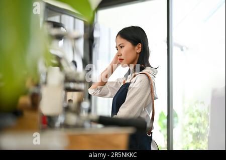 Fokussierte junge asiatische Kassiererin oder Barista, die an der Bar steht und Bestellungen von Gästen entgegennimmt. Café-Bar, Personal, Café-Besitzer, Geschäftsreisender Stockfoto