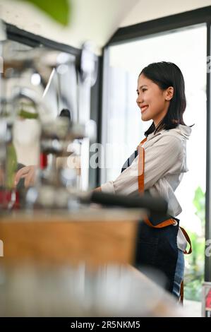 Rückansicht einer freundlichen und lächelnden jungen asiatischen Kassiererin oder Barista, die an der Bar steht und Bestellungen von Gästen entgegennimmt. Gastgewerbe Stockfoto