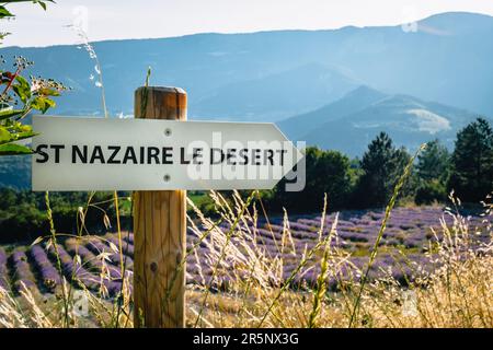 Hölzernes Schild, das auf das kleine Dorf Saint Nazaire le Desert in Drome (Frankreich) hinweist, mit einem Lavendelfeld im Hintergrund Stockfoto