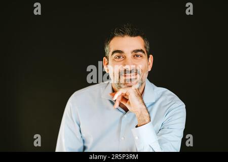 Porträt eines gutaussehenden Mannes, der ein blaues Hemd trägt, sich an eine Hand lehnt, auf schwarzem Hintergrund posiert Stockfoto