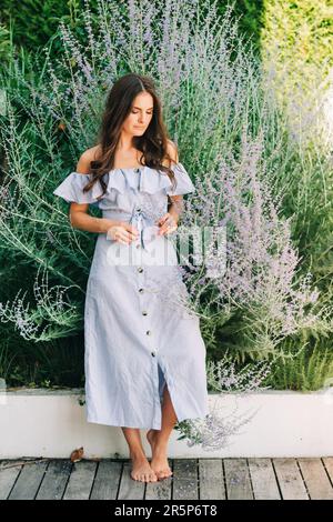 Außenporträt einer hübschen jungen Frau mit dunklem Haar, die im Sommergarten posiert, lila Blumen trägt, blaues Kleid mit offenen Schultern trägt Stockfoto