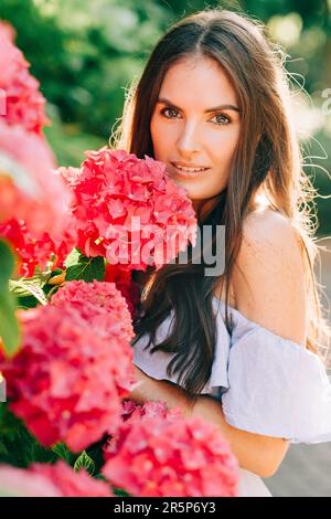 Außenporträt einer schönen Frau, die in rosa Hortensien posiert und ein blaues Kleid trägt Stockfoto