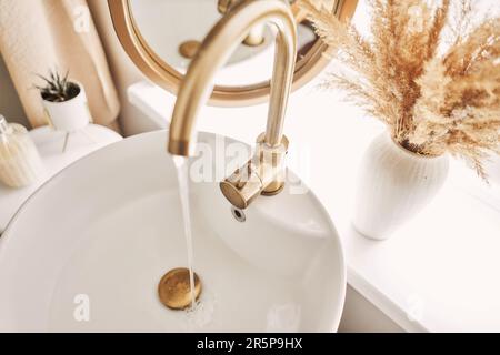 Ein wunderschönes Waschbecken mit angeschaltetem Wasser, goldenem Wasserhahn neben einem ovalen Spiegel und einem Regal mit Handtüchern. Nahaufnahme eines eleganten goldenen Wasserhahns Stockfoto