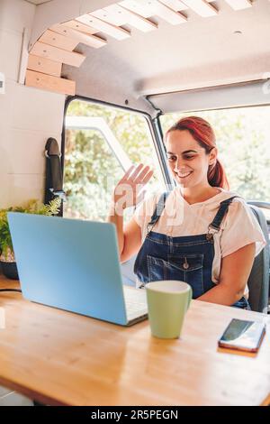 Eine junge Frau, die ein Videogespräch auf dem Laptop in ihrem Wohnwagen hat. Roadtrip im Minibus und Sommerabenteuer im Freien. Nomad Lifestyle-Konzept Stockfoto